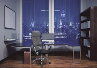 A modern office features a sleek Silla Economalla black chair, a glass desk with a computer and monitor, large windows showcasing a nighttime city skyline with illuminated skyscrapers, along with shelves and a whiteboard decorating the room.