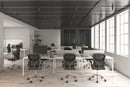 A modern office space features several empty workstations with black Silla Brescia mesh chairs and computer screens. Large windows on the right flood the room with natural light, while a plant is visible in the back corner.